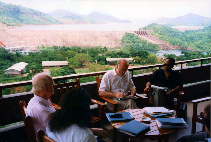 Shireen Kadivar, Amb. John McDonald, Chris Mitchell, and Wallace Warfield sit around a table with various folders and books on a balcony overlooking Akosombo Dam in Ghana.