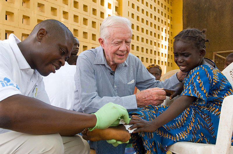 Jimmy Carter holding the hand of a distressed child in Ghana while a health care worker administers care.